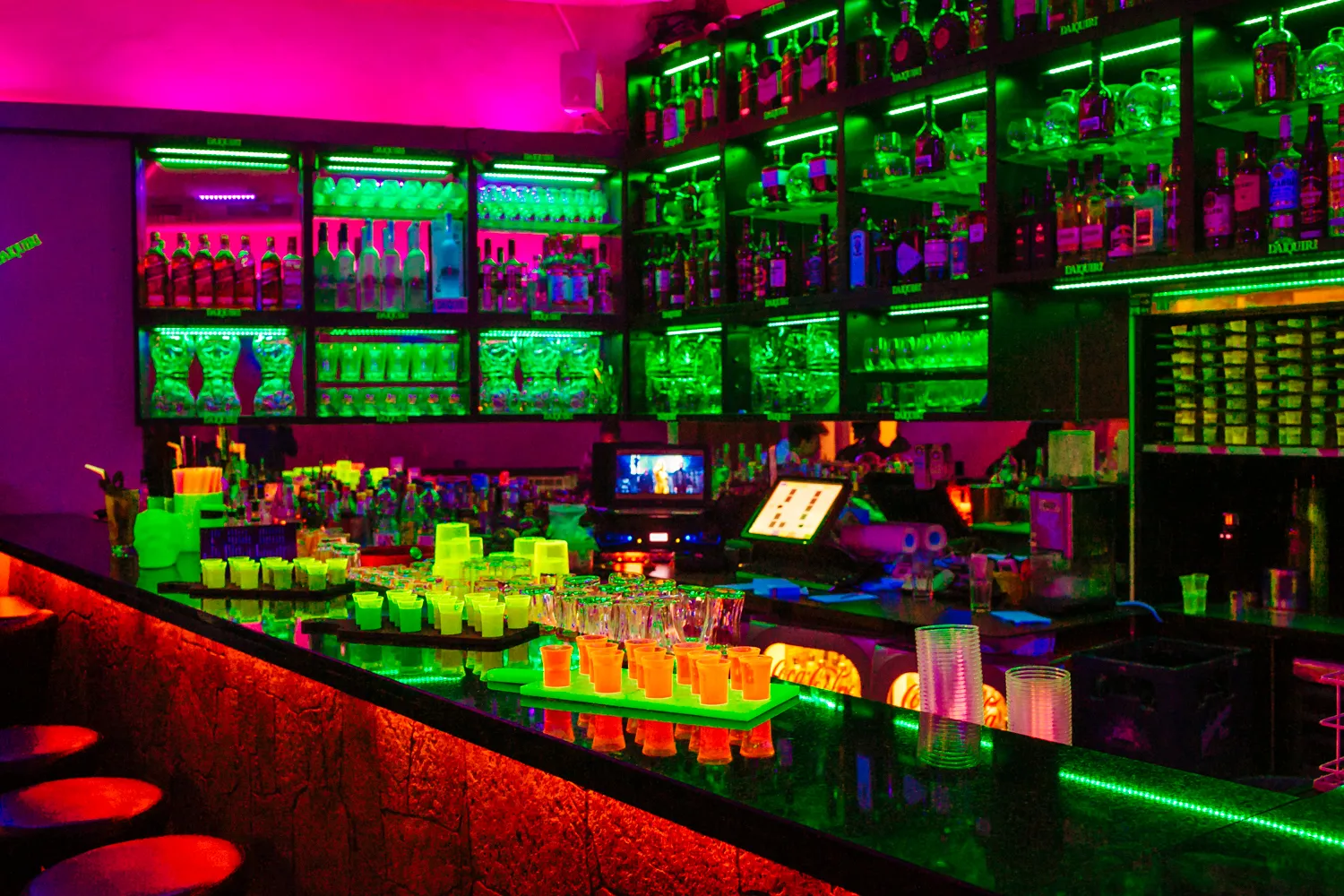 Die Bar unter Neonlicht: beleuchtete Flaschenregale in Pink und Grün, darunter Reihen leuchtender Shots